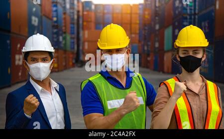 I lavoratori industriali o i tecnici che indossano maschere di protezione Coronavirus o COVID-19 sollevano il pugno e si posizionano davanti al contenitore di spedizione. Foto Stock