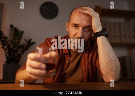 Ritratto di triste uomo maturo seduto al tavolo e guardando la macchina fotografica con bevanda alcolica Foto Stock