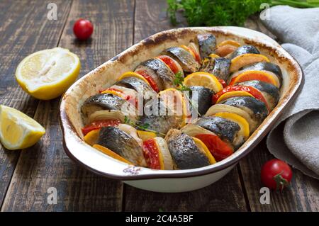 Pesce al forno con verdure. Pezzi di aringhe, cipolle, pomodoro, limone, erbe aromatiche in ceramica su un tavolo rustico in legno Foto Stock