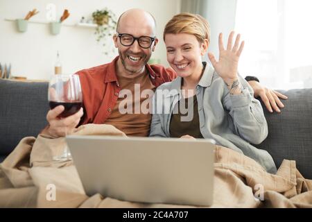 Giovane coppia felice bevendo vino rosso e comunicando online con i suoi amici a casa Foto Stock
