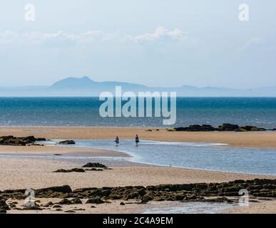 Aberlady, East Lothian, Scozia, Regno Unito, 25 giugno 2020. Tempo in Gran Bretagna: Sole caldo nella baia di Aberlady mentre una coppia si rinfresca in acqua camminando sulla spiaggia a bassa marea con il profilo distintivo e frizzante dello skyline di Edimburgo e Arthur's Seat in lontananza attraverso il Firth of Forth Foto Stock