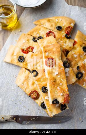 Focaccia fatta in casa con olive nere, pomodori ciliegini, rosmarino, sale di fiocchi e olio d'oliva su carta da forno. Vista dall'alto Foto Stock