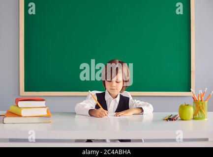 Ritorno a scuola. Divertente ragazzo con capelli lunghi scrive in un notebook al tavolo su uno sfondo di scuola verde lavagna Foto Stock