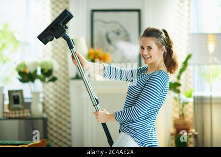 giovane donna sorridente in t-shirt a righe e pantaloni bianchi con aspirapolvere nella casa moderna in giornata di sole. Foto Stock