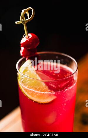 Un cocktail rosso in un bicchiere di highball guarnito con limone e ciliegie Foto Stock