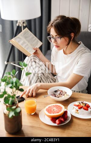 Foto di giovane donna che legge libro e usa il cellulare mentre si ha la colazione in una stanza accogliente a casa Foto Stock