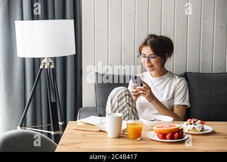 Foto di allegra donna caucasica che usa il telefono cellulare mentre si serve la colazione in una camera accogliente a casa Foto Stock