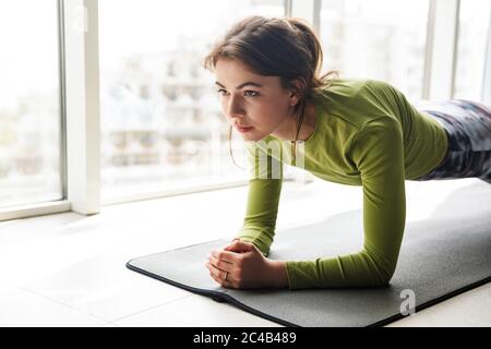 Foto di una donna con bruna focalizzata che fa esercizi di yoga sul tappetino mentre si lavora in una stanza accogliente Foto Stock