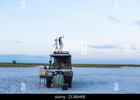 Un fratello e una sorella che si trovano in cima a un veicolo safari al tramonto in un paesaggio di una padella di sale Foto Stock