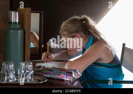 Una ragazza di dodici anni seduta su una scrivania in una tenda in un campo di riserva naturale, disegnando. Foto Stock