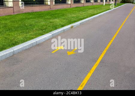 una strada asfaltata con segni gialli e frecce direzionali, una strada con un prato verde e un recinto di pietra. Foto Stock