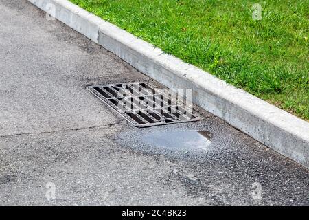 griglia di tombino del sistema di drenaggio sulla strada asfaltata con una puddle dopo la pioggia. Foto Stock