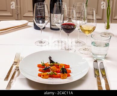 Un antipasto di fette di pesce crudo (forse salmone) con pomodorini gialli tagliati a metà, scaglie pallide (forse ravanello), erbe aromatiche e patatine scure croccanti è disposto su un piatto bianco all’Hotel e Restaurant Kronenschlösschen di Hattenheim, Eltville am Rhein, Assia, Germania. Dietro ci sono bicchieri di vino rosso e bianco, una bottiglia di Notte a San Martino 2013 di Famiglia Olivini e una bottiglia di Riesling Erbach Siegelsberg 2013, Grosses Gewächs (grand cru), dell’azienda vinicola Jakob Jung del Rheingau. Notte a San Martino è un vino rosso con la denominazione Benaco Bresciano del lago di Garda. La degustazione del vino è par Foto Stock