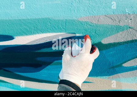 primo piano della mano di un uomo in un guanto tiene un palloncino con vernice e dipinge il muro in blu e bianco Foto Stock