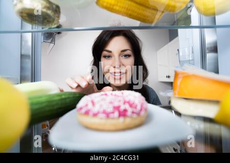 Happy giovane donna che prende Donut da frigorifero o congelatore Foto Stock