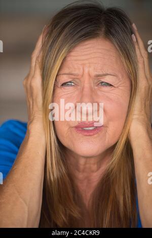 Ritratto attraente donna matura con stressata, dolorosa espressione facciale, mani che coprono le orecchie da rumore forte e suono, sfondo sfocato. Foto Stock