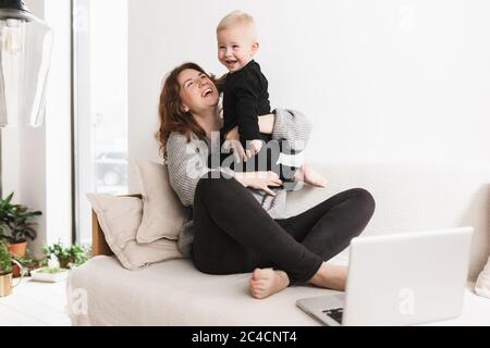 Giovane donna allegra seduta sul divano con il suo piccolo figlio ridente che gioca felice insieme. Mamma e bambino trascorrono con gioia il tempo con il computer portatile Foto Stock