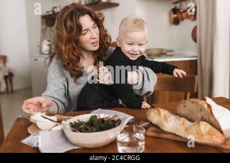 Giovane bella donna con capelli rossi in maglia maglione seduta al tavolo con cibo da mangiare sognante il suo bambino sorridente. Mamma spendendo felicemente Foto Stock