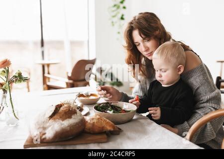 Giovane bella mamma con capelli rossi in maglia maglione seduta al tavolo dreamily nutrire il suo bambino passare il tempo in cucina a casa Foto Stock