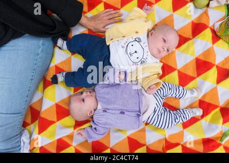 Una madre con i gemelli del bambino e del ragazzo del neonato che si stendano su un tappeto colorato da picnic. La ragazza indossa un cardigan viola e il ragazzo un cardigan giallo. P Foto Stock