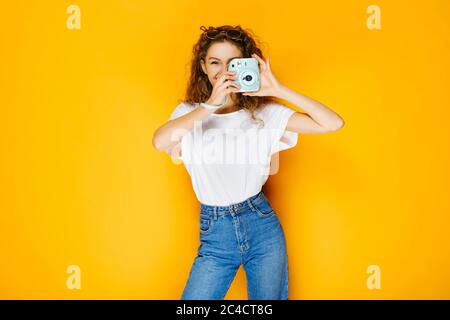 Bella ragazza con fotocamera istantanea isolata su sfondo giallo. Modello che indossa camicia bianca casual e ha lunghi capelli ricci. Foto Stock