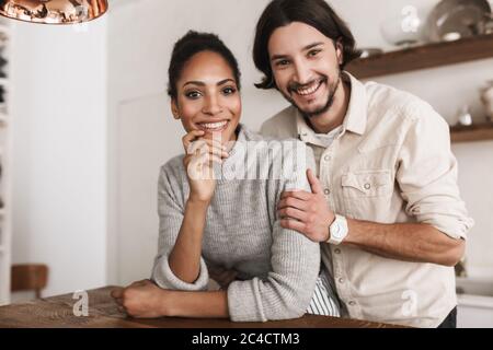 Attraente uomo sorridente e bella donna afroamericana appoggiata sul tavolo gioiosamente guardando in macchina fotografica insieme. Giovane coppia internazionale felicemente Foto Stock