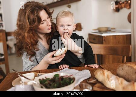 Giovane bella donna con capelli rossi in maglia maglione seduta al tavolo con cibo da mangiare sognando suo figlio. Mamma sognando di passare il tempo con Foto Stock