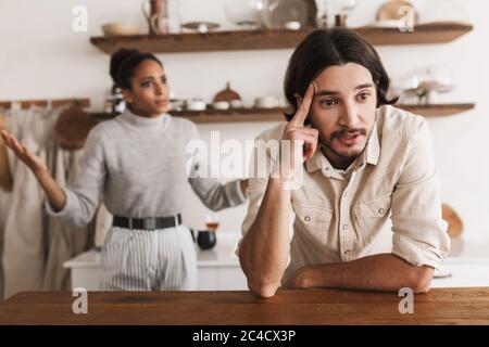 Uomo stanco con barba appoggiata sul tavolo, mentre tristemente guardando da parte con emotiva afroamericana donna sullo sfondo. Giovane coppia internazionale dentro Foto Stock