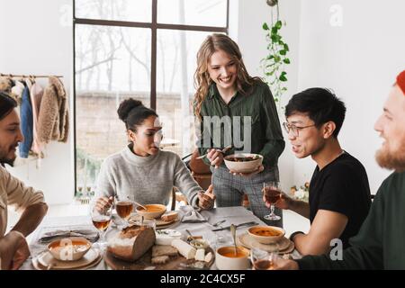 Donna allegra che tiene il ciotola con l'insalata in mano felice di passare il tempo con i colleghi. Gruppo di amici internazionali sedendo al tavolo Foto Stock