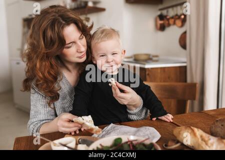 Giovane bella donna con capelli rossi in maglia maglione nutrire il suo piccolo figlio sorridente che gioiosamente guardando in macchina fotografica. Mamma sognando di passare il tempo con Foto Stock