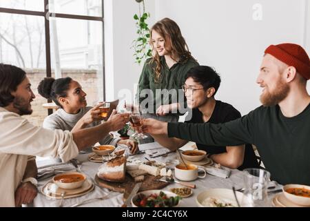 Bella donna con capelli ondulati in piedi vicino a tavola sognando di passare il tempo con i colleghi. Gruppo di giovani amici internazionali che tengono felicemente i bicchieri Foto Stock