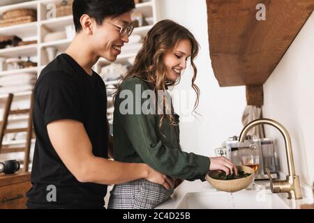 Bella donna felicemente lavare lattuga asiatico uomo dietro abbracciarla. Giovane coppia internazionale sognando di passare il tempo insieme in cucina a casa Foto Stock