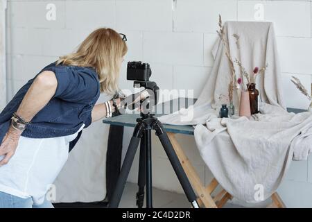 Vista posteriore di una donna creativa impegnata che utilizza la fotocamera sul treppiede mentre fotografa la composizione del vaso in studio Foto Stock