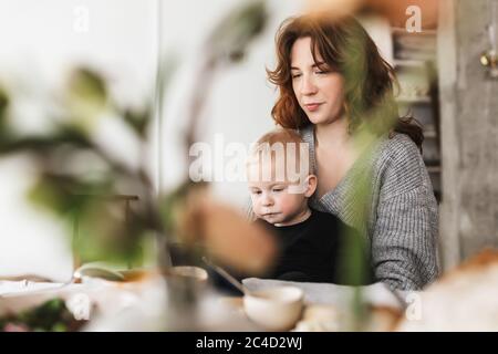 Giovane bella donna con capelli rossi in maglia maglione seduta al tavolo dreamily nutrendo il suo bambino. Mamma che passa il tempo con il bambino Foto Stock