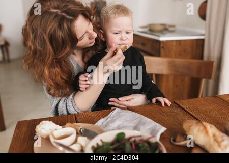 Giovane donna attraente con capelli rossi in maglia maglione seduta al tavolo con cibo che nutrono pensieroso il suo bambino. Mamma che passa il tempo con Foto Stock