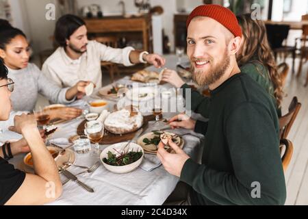 Bell'uomo sorridente in cappello rosso lavorato a maglia con barba seduta al tavolo che guarda felicemente in macchina fotografica. Gruppo di giovani amici internazionali che trascorrono del tempo Foto Stock
