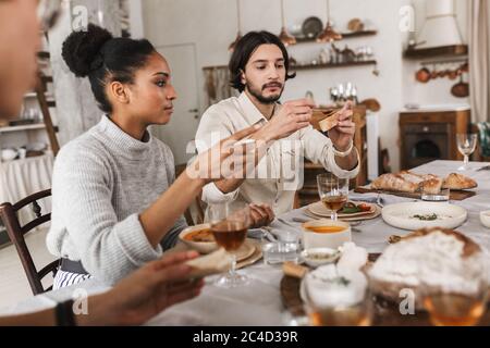 Bella donna americana afro e bell'uomo con barba seduto al tavolo da mangiare pane sognante. Gruppo di giovani amici internazionali che trascorrono del tempo Foto Stock