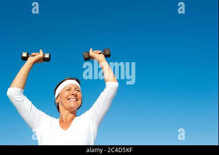 Donna anziana attraente e attiva che si esercita con pesi, isolata con cielo blu come sfondo e spazio di copia. Foto Stock