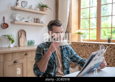 Uomo alto in una camicia a scacchi seduto al tavolo, con caffè e leggendo un giornale Foto Stock
