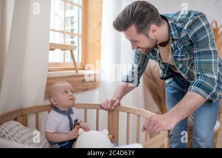 Padre dai capelli scuri che sta vicino a playpen guardando suo figlio piccolo Foto Stock