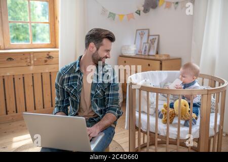 Padre dai capelli scuri che lavora su un computer portatile e sorridendo mentre suo figlio gioca Foto Stock
