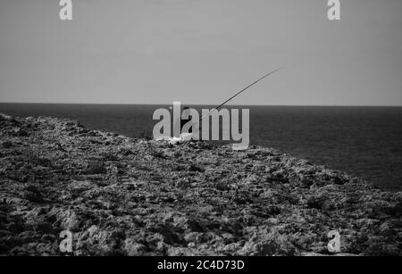 MELLIEHA, MALTA - 12 ottobre 2014: Un uomo che pesca da solo in pace su una scogliera a Malta, guardando verso l'orizzonte, seduto sul paesaggio carsico pietra calcarea ro Foto Stock