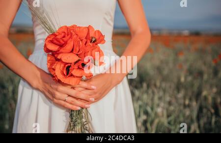 Sposa in un abito bianco che tiene un bouquet di fiori di papavero, caldo tempo di tramonto sullo sfondo del campo di papavero rosso. Spazio di copia. Il concetto di Foto Stock