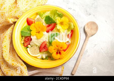 insalata di fiori di nasturzio con pomodori e limoni in ciotola di ceramica gialla. Cucchiaio di legno, cibo vegetariano. Foto Stock