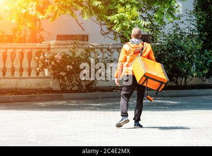 Uomo di consegna con zaino. Deliveryman caucasico isolato in uniforme arancione su sfondo parco. Servizio di consegna senza contatto durante il corso Foto Stock