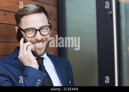 Uomo d'affari maturo di successo in occhiali e in tuta blu che parla al telefono cellulare all'aperto Foto Stock