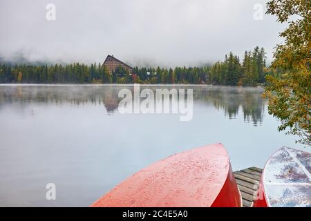 Lago di montagna Strbske Pleso con sfondo foggoso e barche rosse capovolte sul molo in primo piano. Foto Stock