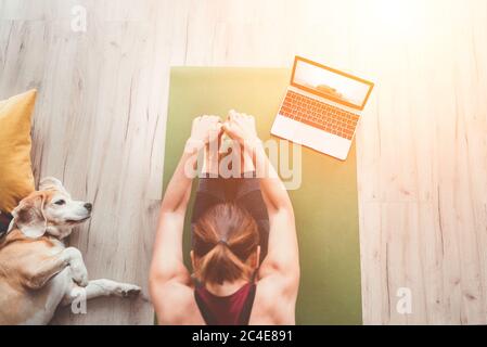 Vista dall'alto in forma sportiva donna sana seduta sul tappetino in posa Paschimottanasana, facendo esercizi di respirazione, guardando una lezione di yoga online sul computer portatile. H Foto Stock