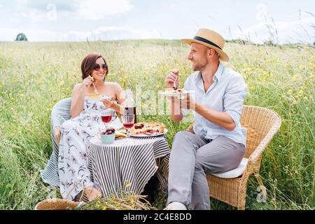 Felice coppia di mezza età seduta in sedie di rattan sul prato d'erba accanto al tavolo servito con torta di frutta e stoviglie vintage, mangiare un ta Foto Stock