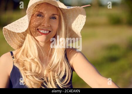 Bella donna bionda sorridente e amichevole con capelli lunghi che indossa un cappello di paglia all'aperto in una giornata estiva in un ritratto ravvicinato Foto Stock